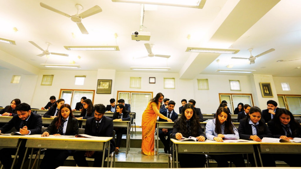 Students in uniforms taking an examination in a well-lit classroom while a teacher monitors.