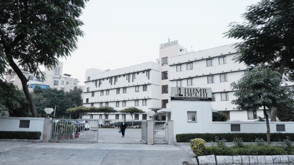 Front view of a modern educational building with a signboard reading "iihmr delhi" flanked by trees and a person walking by the gate.